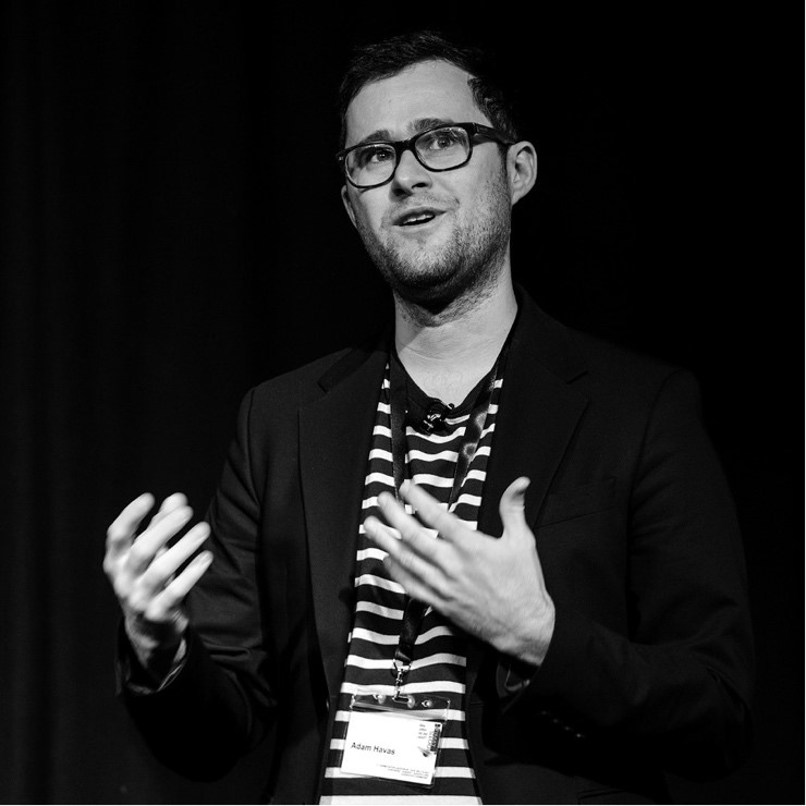 Black and white image of a man with short hair, wearing dark framed glasses, with close shaven beard. Wearing blazer and striped shirt in midst of talk and gesturing with hands.